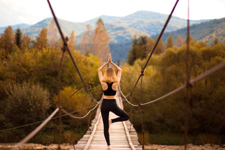 Young woman standing in tree yoga pose, meditating on bridge over mountain river. Zen, meditation, peaceの写真素材