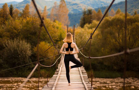 Young woman standing in tree yoga pose, meditating on bridge over mountain river. Zen, meditation, peaceの写真素材