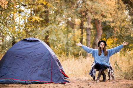Young woman tourist sitting near tent in blue sweater and gray hat and enjoy freedom at autumn forest sunsetの写真素材