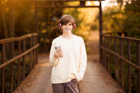 Young beautiful teen girl in headphones listening to music in phone while walk by street at autumn sunsetの写真素材