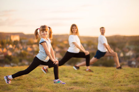 Parents instill sports habits in their child by example, focus on child. Happy sporty family doing stretching exercises, practices yoga outdoor. Mom dad and daughter doing sport exercises together.の写真素材