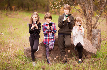 Group of school children playing in mobile phone together while standing outdoors in summer park
