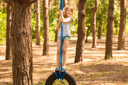 Cute little girl swinging on wheel attached to big tree in autumn forest.の写真素材