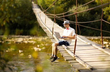 Concentrated young businessman in eyewear sit on bridge across mountains river with laptop. Web surfing information in internet or working distantly online. Communication remotely with client or study.の写真素材