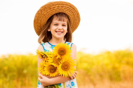 Outdoor portrait of adorable little girl in straw hat and blue dress is walking in a field of sunflowers, gathering and collecting a bouquet. Summer holidaysの写真素材