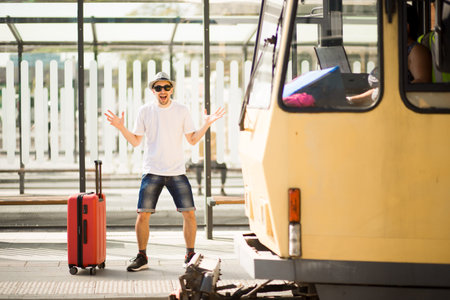 Young traveller man in hat and sunglasses with suitcase running by train station. Tourist late on...