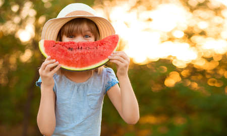 Shy little girl in hat hold big slice of watermelon and hide her face outdoor. Summer holidays full of vitaminの写真素材