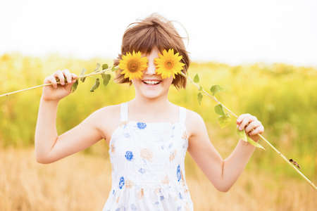 Cute child girl wear dress with sunflower in summer field. Happy little girl hide eye with sunflower. Summer time concept.の写真素材