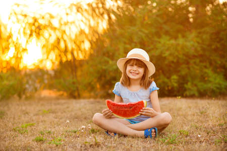 Adorable baby girl in straw hat sit with cross legs on green grass, and eagerly eats juicy watermelon with summer sunshine on backgroundの写真素材