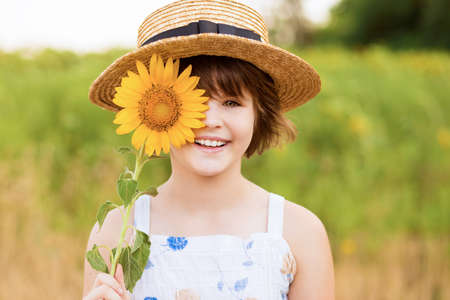 Beautiful little girl in straw hat with fluttering hair smile and hide eye with sunflower flower, walking outdoor in summer holidayの写真素材