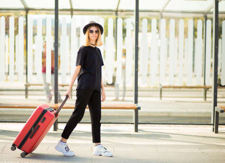 Young casual female traveler in total black and sunglasses walking and pulling a red wheeled luggage. Concept of travel. Copy spaceの写真素材
