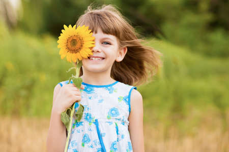 Cute child girl wear dress with sunflower in summer field. Happy little girl hide eye with sunflower. Summer time concept.の写真素材