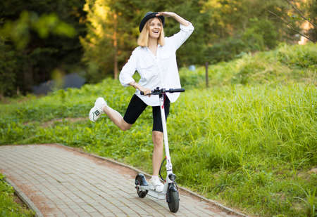 Cheerful beautiful girl in black hat and white shirt riding an electric scooter in urban style in summer park. Scooter rental, freedom of movementの写真素材
