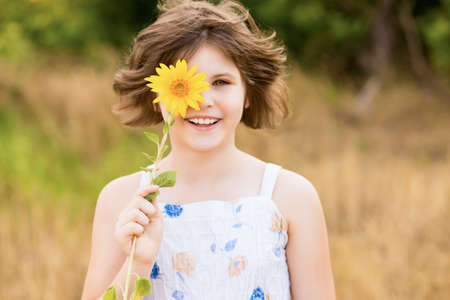 Cute child girl wear dress with sunflower in summer field. Happy little girl hide eye with sunflower. Summer time concept.の写真素材