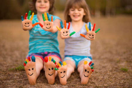 Two funny child girls sitting on grass and waving painted hands and foot in funny faces in summerの写真素材