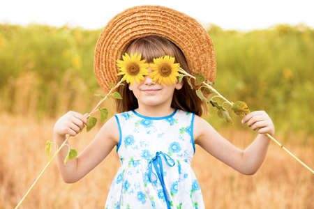 Beautiful little girl in straw hat with fluttering hair smile and hide eye with sunflower flower, walking outdoor in summer holidayの写真素材