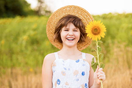 Beautiful little girl in straw hat with fluttering hair smile with sunflower flower, walking outdoor in summer holidayの写真素材