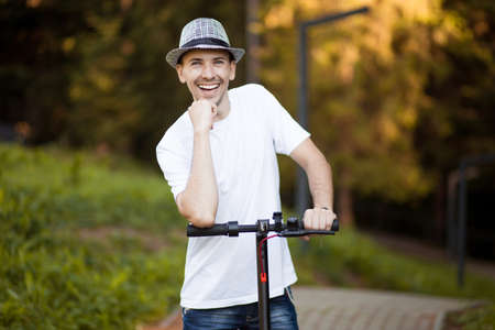 Portrait of young handsome man in white shirt, sunglasses and hat ready to ride his electric scooter bike on city street in sunny summer. Ecological transport concept. Electric urban transportation.の写真素材
