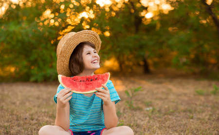 Adorable baby girl in straw hat sit with cross legs on green grass, and eagerly eats juicy watermelon with summer sunshine on backgroundの写真素材