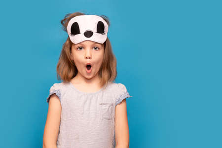 Portrait of young girl with surprised face and sleeping mask on head isolated over blue background. Empty space for textの写真素材