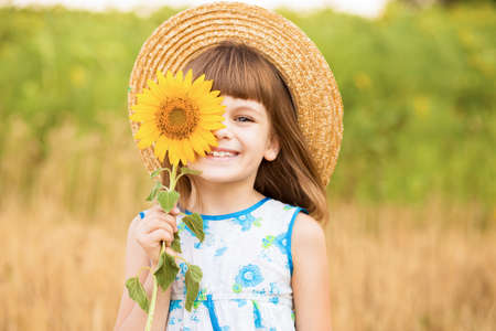 Beautiful little girl in straw hat with fluttering hair smile and hide eye with sunflower flower, walking outdoor in summer holidayの写真素材