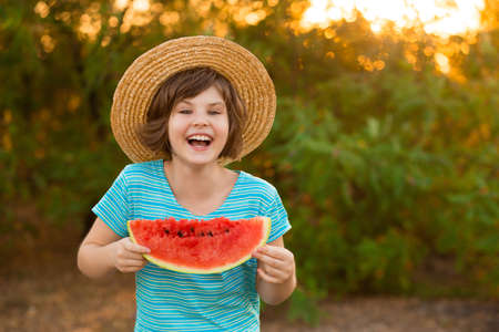 Adorable baby girl in straw hat smile and eagerly eats juicy watermelon with summer sunshine on backgroundの写真素材