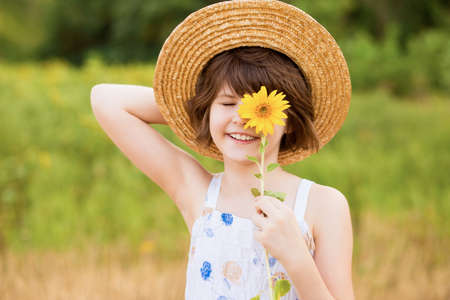 Beautiful little girl in straw hat with fluttering hair smile and hide eye with sunflower flower, walking outdoor in summer holidayの写真素材