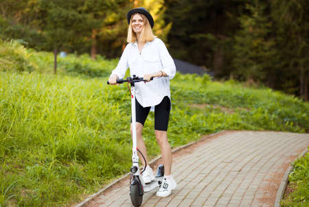 Cheerful beautiful girl in black hat and white shirt riding an electric scooter in urban style in summer park. Scooter rental, freedom of movementの写真素材