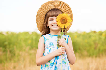 Beautiful little girl in straw hat with fluttering hair smile and hide eye with sunflower flower, walking outdoor in summer holidayの写真素材