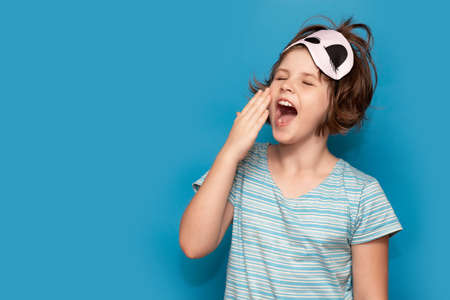Photo of adorable sweet kid nightwear yawning stretch. Sleepy child girl isolated on blue background yawns, sleep mask on her head. Banner design. Copy spaceの写真素材