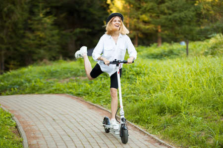 Cheerful beautiful girl in black hat and white shirt riding an electric scooter in urban style in summer park. Scooter rental, freedom of movementの写真素材