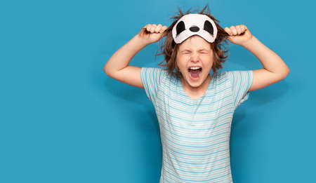 Spoiled little girl in sleeping mask, tearing her hair with closed eyes isolated on blue background. Stressed kid. Screaming, be mad and naughty. Angry child want sleep. Genuine human emotions, reaction and body language.の写真素材