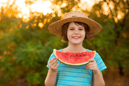 Adorable baby girl in straw hat smile and eagerly eats juicy watermelon with summer sunshine on backgroundの写真素材