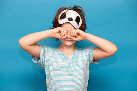Portrait of tired girl with sleeping mask cry and rub her eyes after sleep in morning isolated over blue background. Female kid does not want to wake up and go to school. Morning concept.の写真素材