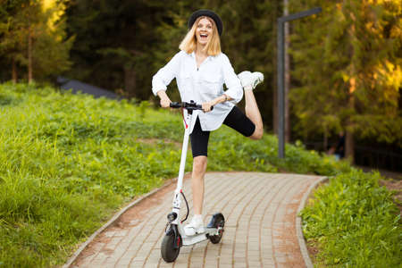 Cheerful beautiful girl in black hat and white shirt riding an electric scooter in urban style in summer park. Scooter rental, freedom of movementの写真素材