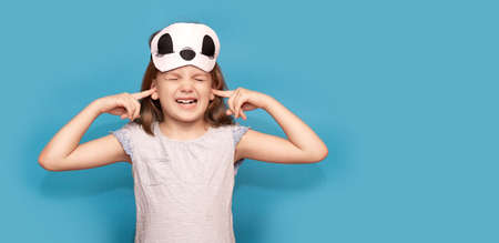 Portrait of stressed child with sleeping mask isolated on blue background. Little girl cover her ears with fingers ask to stop noiseの写真素材