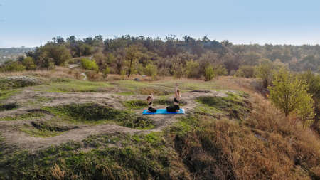 Aerial view of mother and daughter sitting on comfy yoga mat in lotus pose and do yoga exercises at sunrise outdoor on top of hill. High quality photoの写真素材