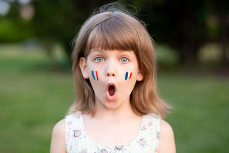 Portrait of surprised 7 years old child with painted cheeks in flag of France walking outdoor. Cute little girl screaming with opened mouth and crazy expression looking at camera.の写真素材
