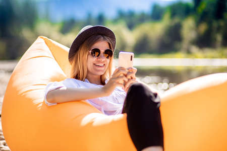 Portrait of charming smiling woman in sunglasses using cellphone and write messages to friends while lying on inflatable mattress on summer beachの写真素材