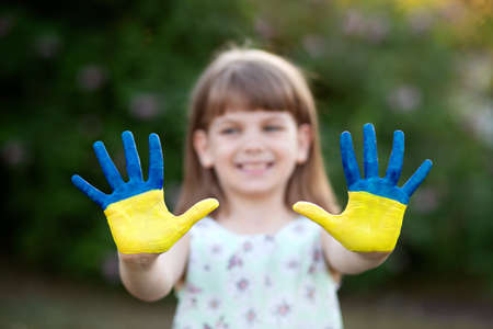 Joyful child girl waving hands painted in Ukraine flag colors and say hello outdoor at nature background, focus on handsの写真素材