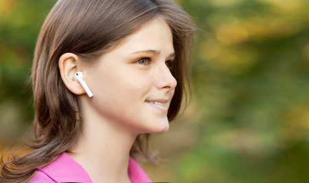 Close up portrait of lovely girl in pink listening to music through wireless earphones outdoors. Young girl smiling happy.の写真素材