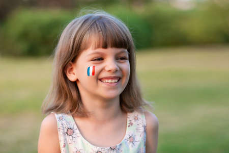 Outdoor portrait of little caucasian kid girl with cheeks painted in France flag colors and looking into camera and smile. Young painter. Modern Art.の写真素材