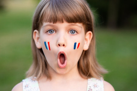Portrait of surprised 7 years old child with painted cheeks in flag of France walking outdoor. Cute little girl screaming with opened mouth and crazy expression looking at camera.の写真素材