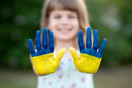 Joyful child girl waving hands painted in Ukraine flag colors and say hello outdoor at nature background, focus on handsの写真素材