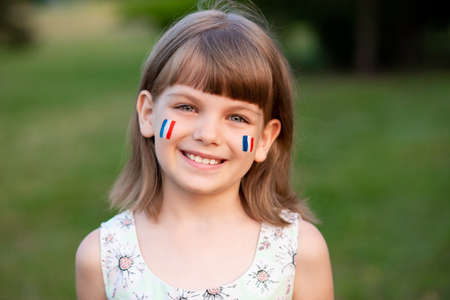 Outdoor portrait of little caucasian kid girl with cheeks painted in France flag colors and looking into camera and smile. Young painter. Modern Art.の写真素材