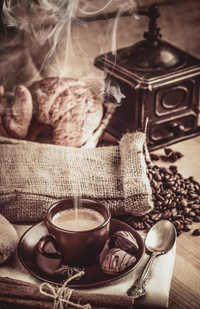 Cup fragrant hot coffee with bean chocolate sweet and croissant in vintage rustic style cinnamon steam smoke on wooden board for bracing morning breakfast.の写真素材