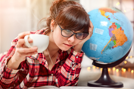 Student girl study homework and reads book. Cup of hot coffee drink in hand. Confused tired face with glasses. Globe world planet Earth.の写真素材