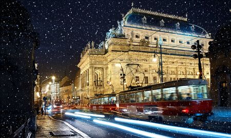 Prague, Czech Republic. Nighttime view to National Theater (Narodni Divadlo) at promenade of river Vltava with bridge, evening illumination and street lamps. Urban winter snowfall landscape with trams.の写真素材