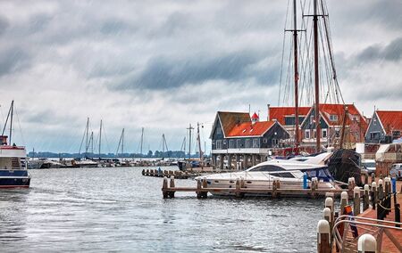 Volendam, Amsterdam, Netherlands. Luxury yachts by piers. Sea bay with traditional dutch houses with red roofs in famous touristic village. Rainy weather with heavy shower rain and dramatic sky clouds.の写真素材