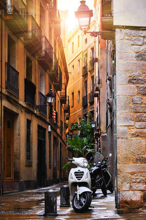 Barcelona, Catalonia, Spain. Romantic narrow residential bystreet in Gothic Quarter. Antique street lamp at wall of old house, parked motorbikes and arch with window between buildings.の写真素材